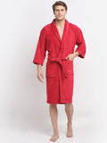 A man wearing a red terry cloth bathrobe with a shawl collar, waist tie, and front patch pockets, standing against a plain light gray studio background.