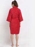 A woman viewed from the back wearing a bright red bathrobe with three-quarter sleeves and a matching waist tie, standing against a plain light gray studio background.