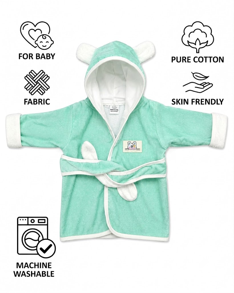 A baby wearing a mint green plush hooded bathrobe with white trim and bear ears on the hood, sitting on a white faux fur rug next to a small red toy car.