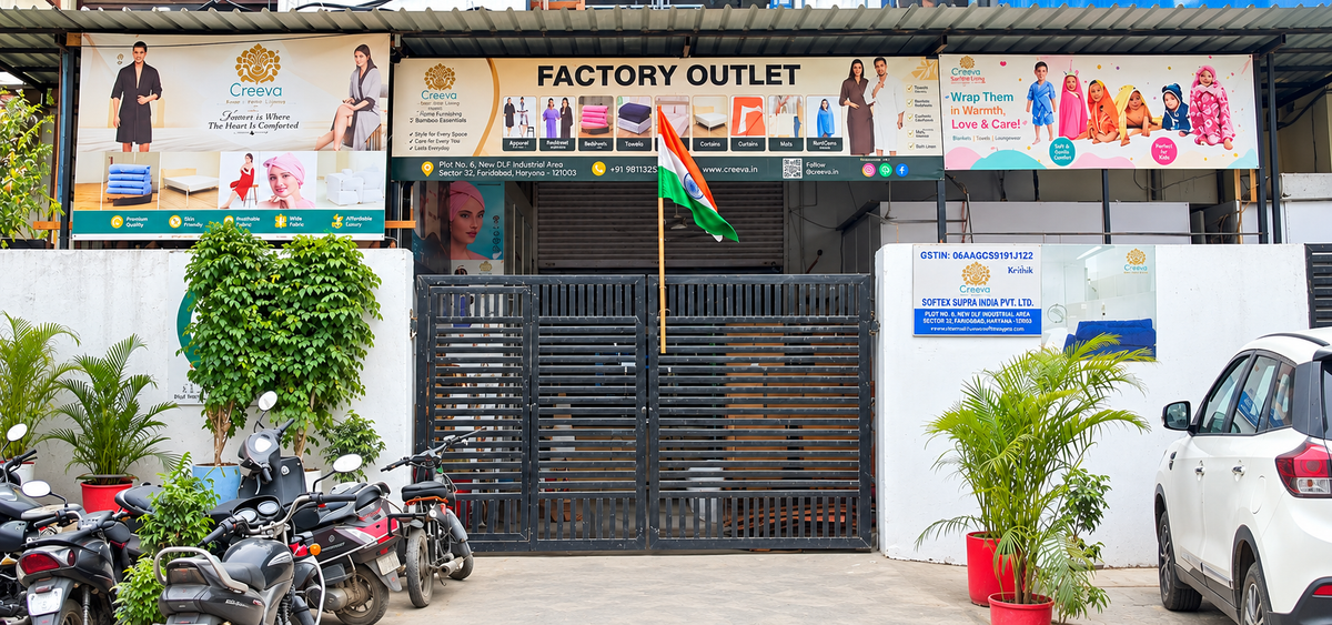 Factory outlet storefront with a black metal gate, large multicolored promotional banners, potted plants, parked motorcycles on the left, and a white car on the right.slider_item_JxCMa9