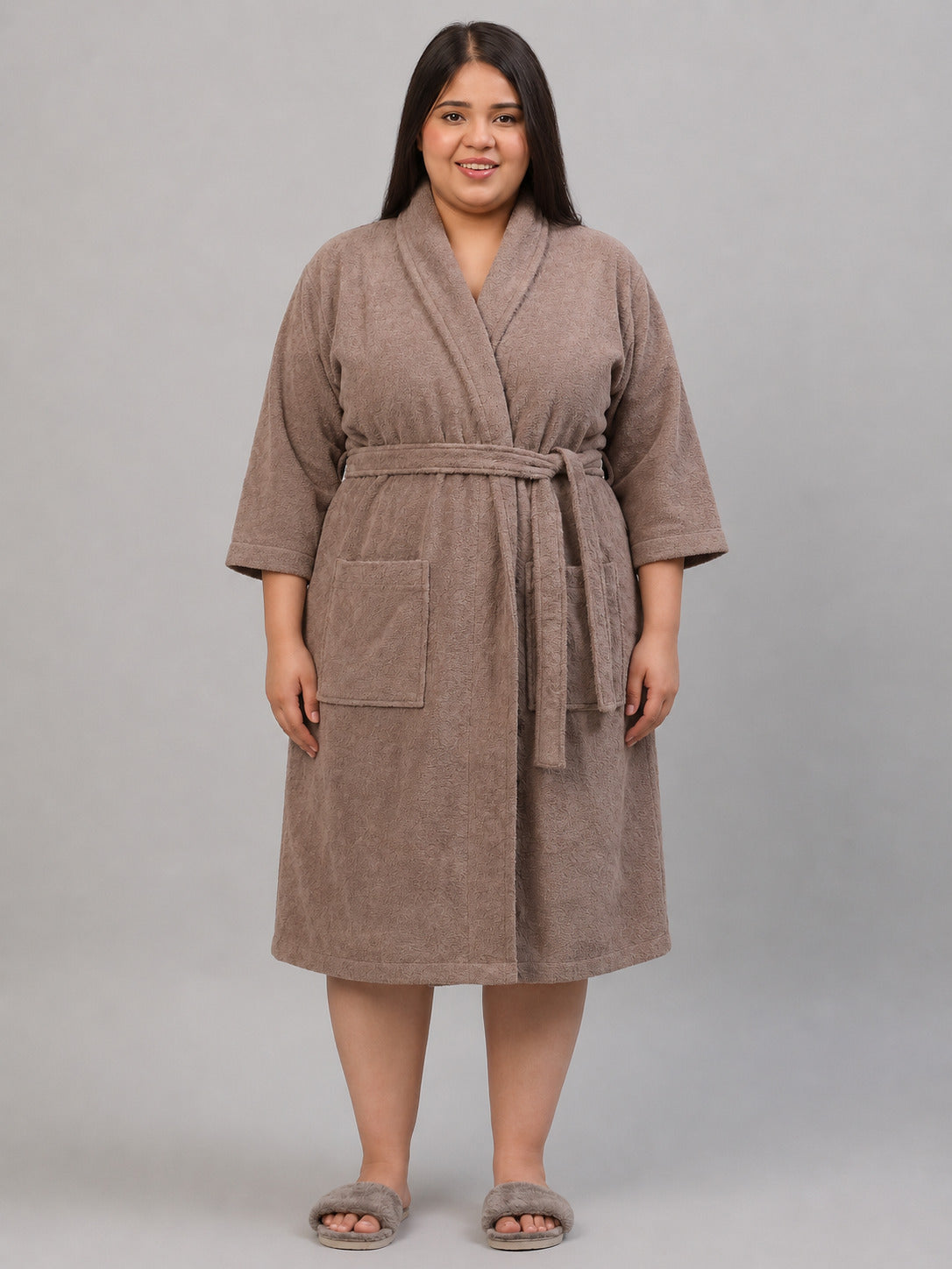 A woman wearing a plush taupe-brown bathrobe stands next to a wooden stool, holding a white mug. The robe features a shawl collar, a matching tie belt, and front patch pockets.