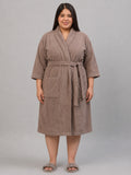 A woman modeling a taupe brown terry cloth bathrobe with a tie belt and front patch pockets, standing against a solid gray studio background.