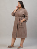 A woman modeling a taupe brown plush bathrobe with a tie waist belt and front patch pockets, paired with matching taupe slippers, against a neutral gray studio background.