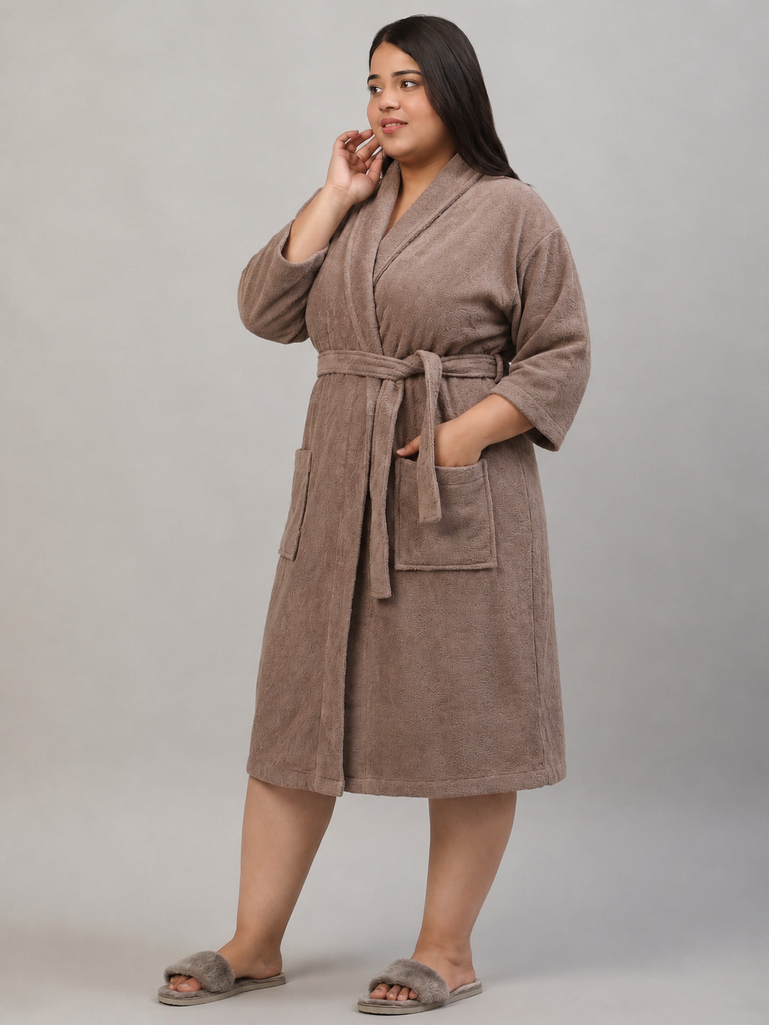 A woman modeling a taupe brown plush bathrobe with a tie waist belt and front patch pockets, paired with matching taupe slippers, against a neutral gray studio background.