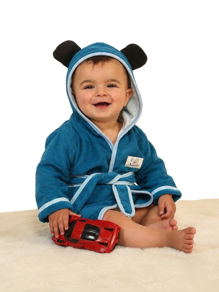 A baby wearing a blue plush hooded bathrobe with black bear ears and white trim, sitting on a cream rug and holding a red toy car.