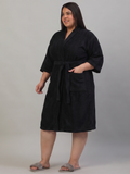A woman modeling a black bathrobe with a tie waist and front pockets, standing against a plain light gray studio background.