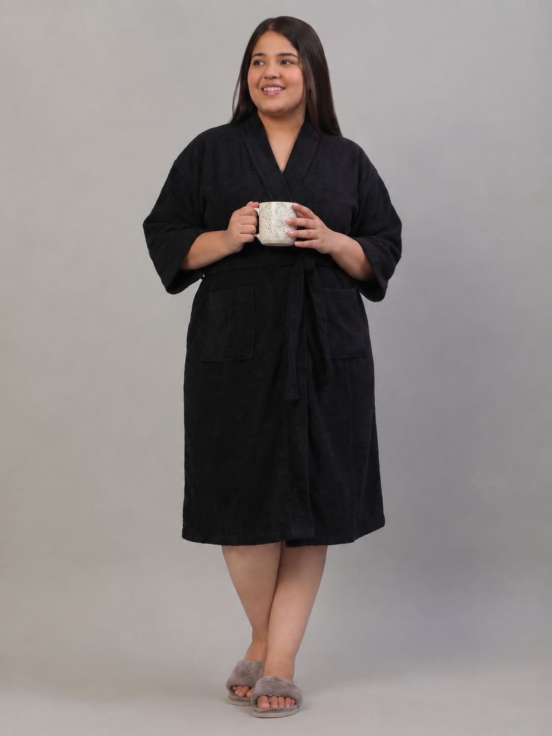 A woman modeling a black cotton bathrobe with a tie belt and front pockets, holding a white mug, against a solid gray studio background.