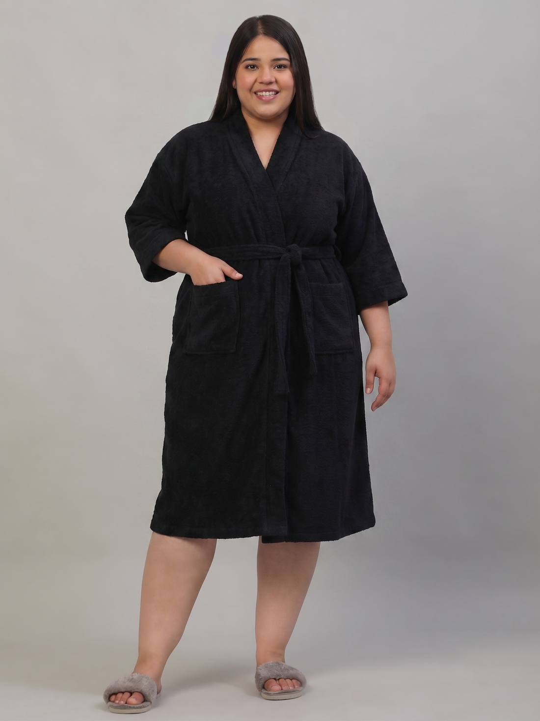 A woman modeling a black cotton bathrobe with a tie belt and front pockets, holding a white mug, against a solid gray studio background.