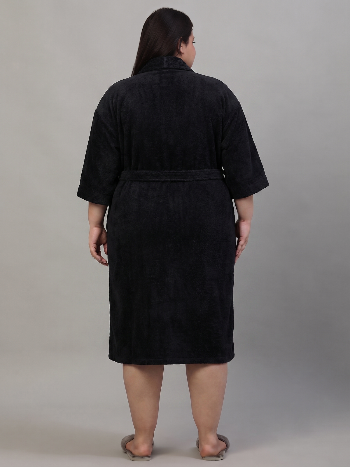 A person shown from the back wearing a black bathrobe with three-quarter sleeves and a tie belt, standing against a plain light gray studio background.
