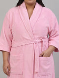 A pink terry cloth bathrobe with a shawl collar, self-tie belt, and front patch pockets, shown on a model against a neutral gray background.