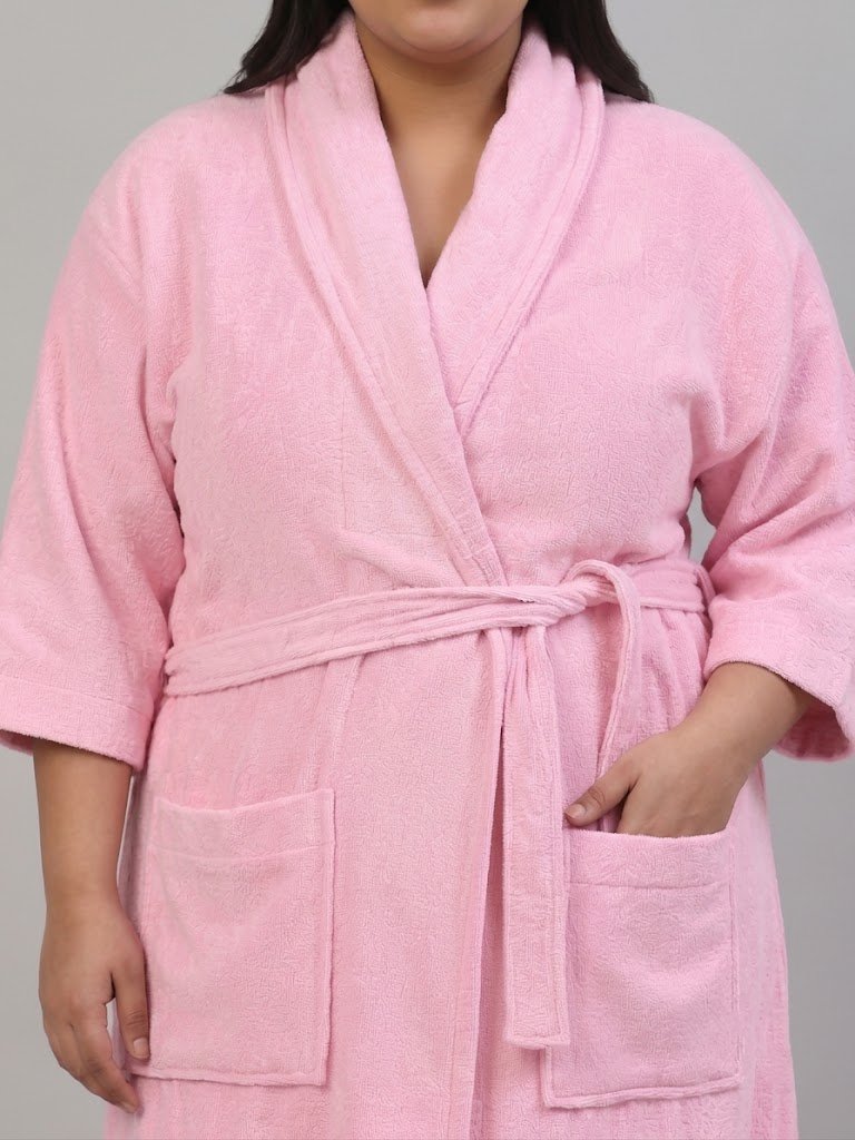 A pink terry cloth bathrobe with a shawl collar, self-tie belt, and front patch pockets, shown on a model against a neutral gray background.