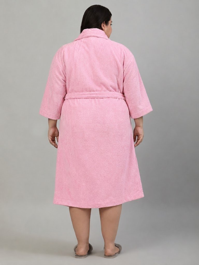 A woman modeling a pink bathrobe with a shawl collar, waist tie, and front patch pockets, standing against a plain gray studio background.