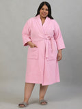 A woman modeling a pink bathrobe with a shawl collar, waist tie, and front patch pockets, standing against a plain gray studio background.
