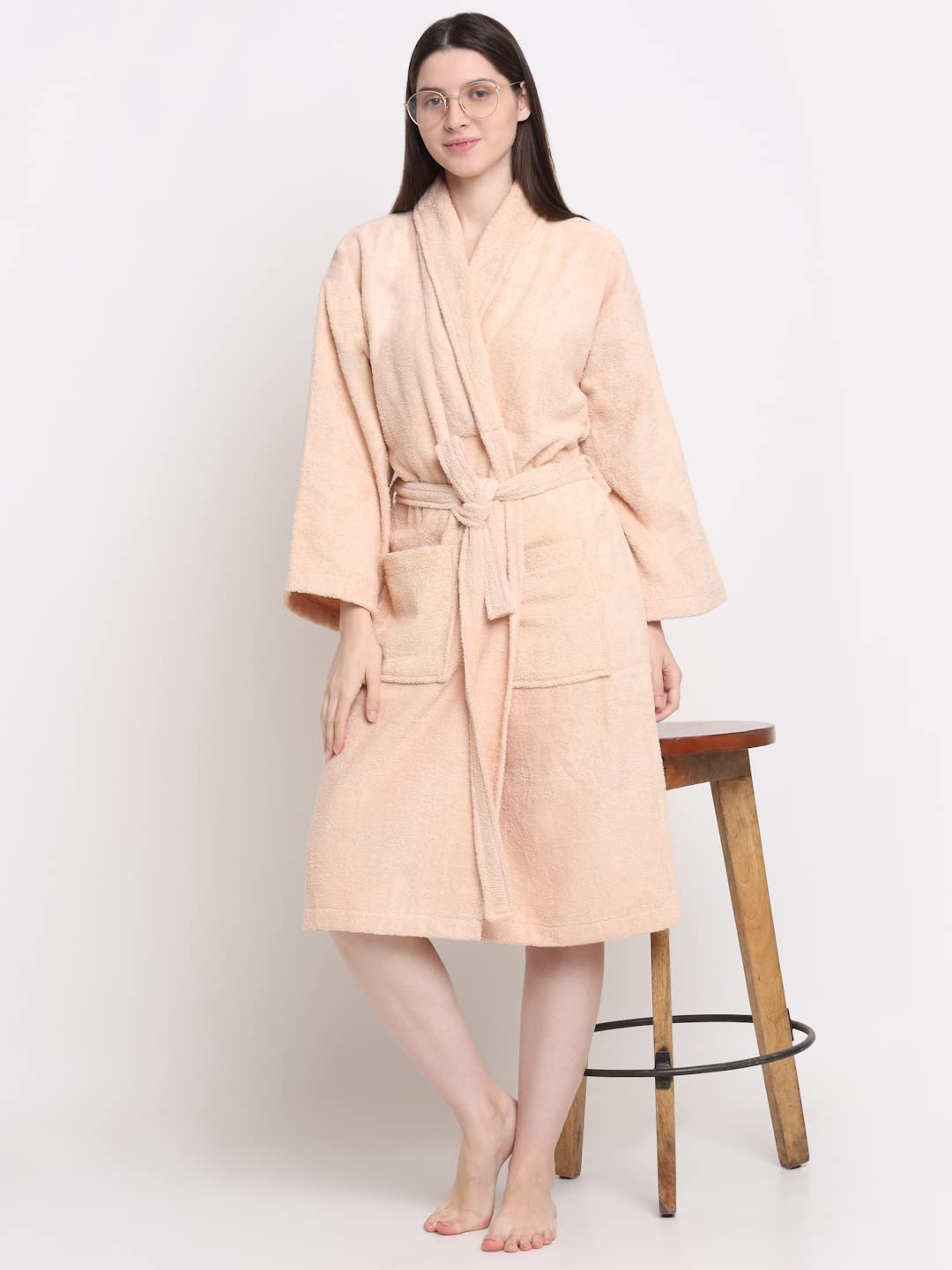 A woman modeling a light peach-colored terry cloth bathrobe with a matching tie belt, standing against a plain white background.