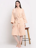 A woman wearing a light peach-colored plush bathrobe stands barefoot next to a wooden stool against a plain white studio background.