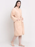 A woman modeling a light peach-colored plush bathrobe with a tie belt and front patch pocket, standing against a plain white studio background.