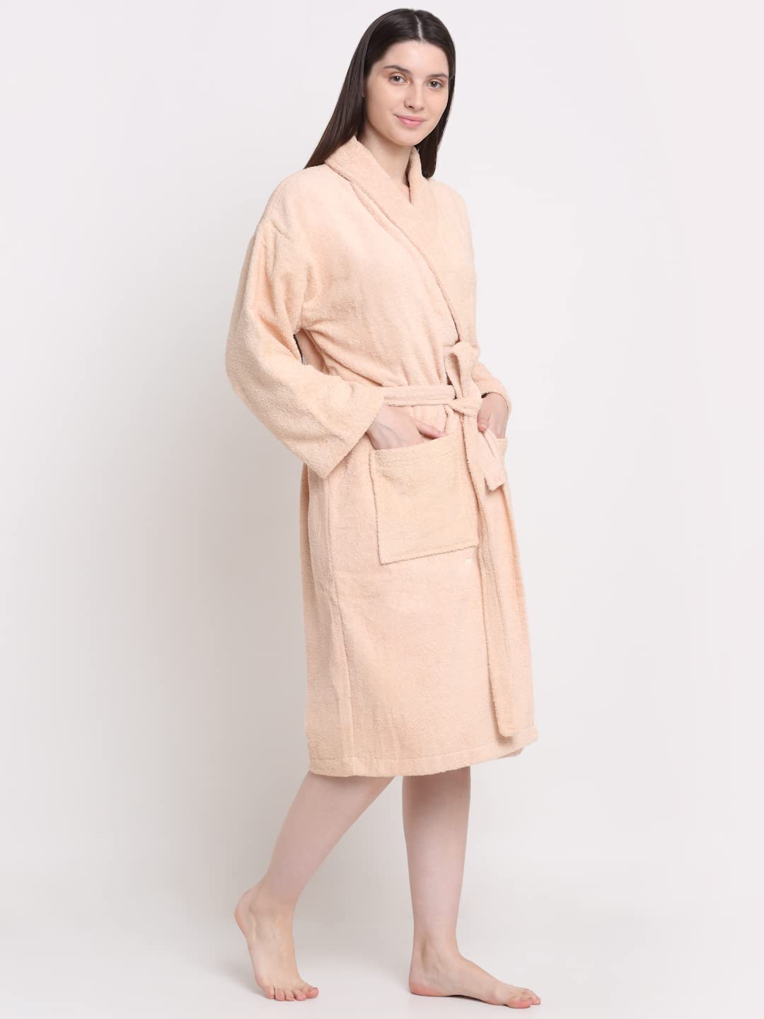 A woman modeling a light peach-colored plush bathrobe with a tie belt and front patch pocket, standing against a plain white studio background.