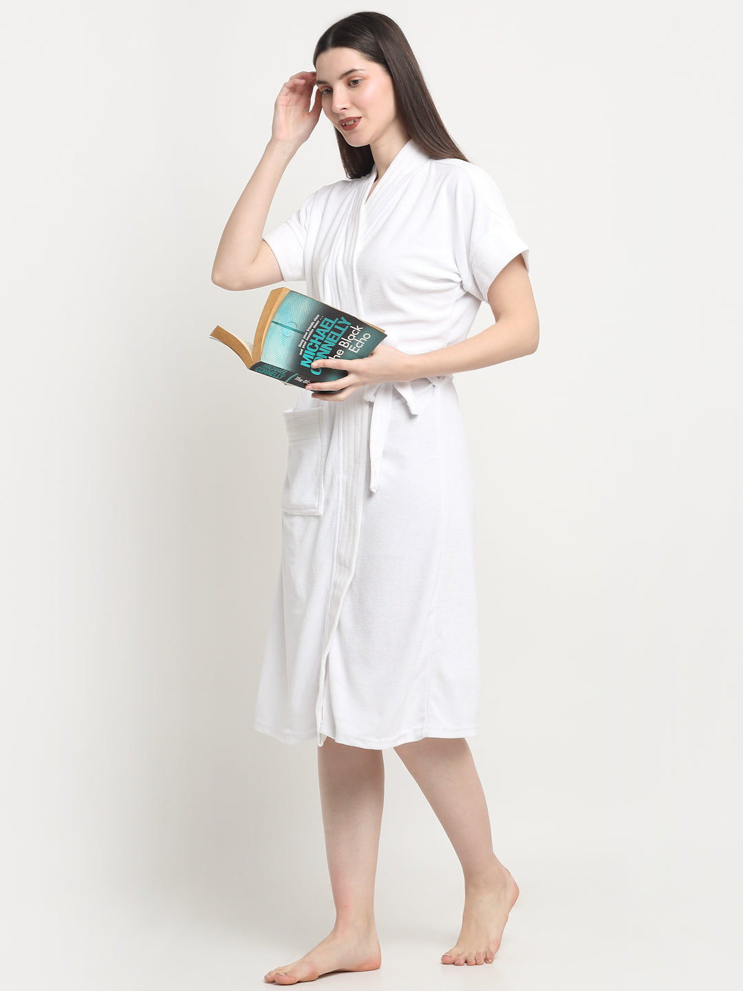 A woman stands wearing a white micro terry bathrobe and holding a book