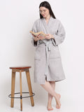A woman standing next to a wooden stool, wearing a silver, hooded, 100% cotton bathrobe with a tie waist, reading a book.