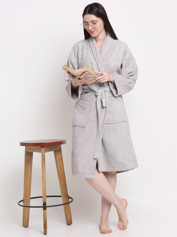 A woman standing next to a wooden stool, wearing a silver, hooded, 100% cotton bathrobe with a tie waist, reading a book.