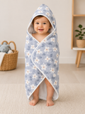 A baby wrapped in a light blue hooded towel with a white and blue floral pattern, standing barefoot on a beige carpet in a bright room.