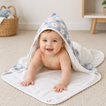 A baby lying on their stomach on a beige carpet, wrapped in a white hooded towel with a light blue and white daisy pattern on the hood and corners.
