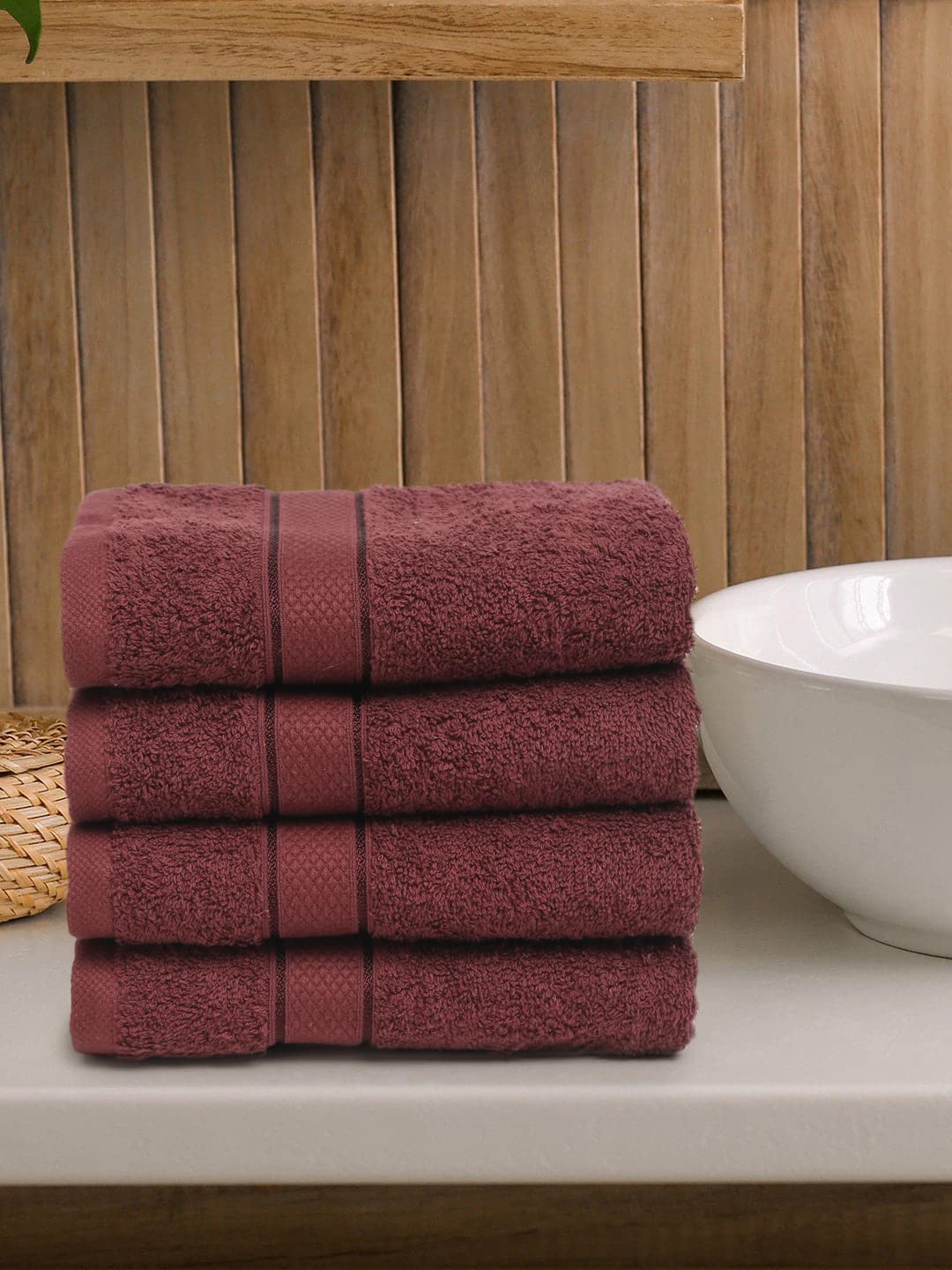A set of four wine-colored Creeva 100% cotton hand towels, neatly stacked, with a wooden backdrop and a white bowl in the foreground.
