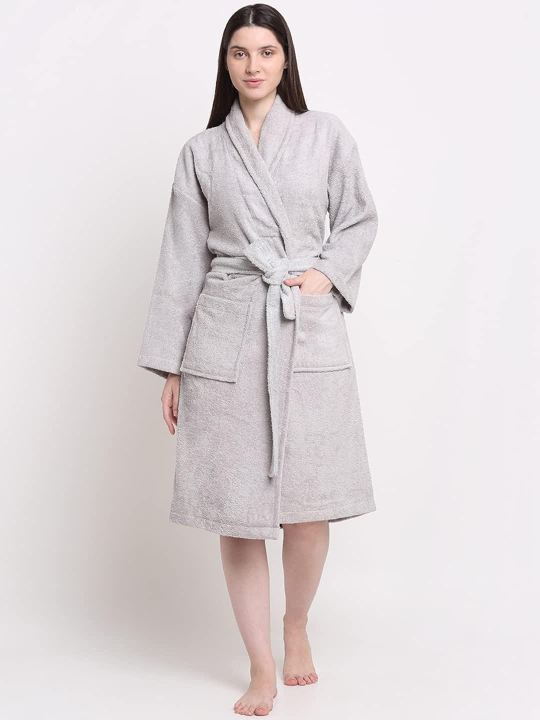 A woman standing next to a wooden stool, wearing a silver, hooded, 100% cotton bathrobe with a tie waist, reading a book.