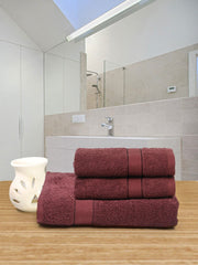Stack of maroon towels on a wooden surface with a white vase, mirrored bathroom backdrop.
 creeva wine color towels